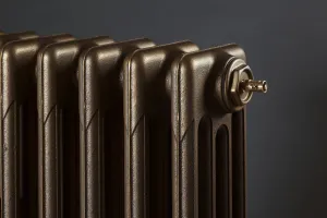 Close-up of the end of a plain Mercury 4 Column cast iron radiator in Beaten Brass with a brass bleed valve, against a dark grey wall.
