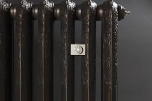 Close-up of an ornate Rococo II cast iron radiator in a dark bronze finish, with a cream wall stay and brass bleed valve, against a grey wall.
