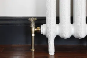 Chatsworth manual angled valve in Antique Brass fitted to an ornate white cast iron radiator on feet, dark navy skirting and a wooden floor.