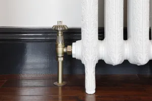 Chatsworth angled valve in Natural Brass on a floor pipe beside an ornate white cast iron radiator on feet, dark skirting and wood floor.