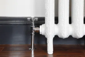 Chrome Crosshead manual angled valve on an ornate white cast iron radiator on feet, against a dark skirting and wooden floor.