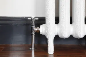 Chrome Crosshead angled manual valve fitted to the foot of an ornate white cast iron radiator, by a dark grey skirting and wooden floor.