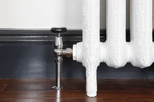 Chrome Windsor angled manual valve with a black head on an ornate white cast iron radiator on feet, by a dark skirting and wooden floor.