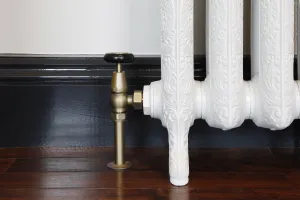 Windsor angled manual valve in Natural Brass with a black wheel head, fitted to an ornate white cast iron radiator on feet by a dark skirting board.