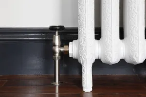 Windsor angled manual valve in Satin Nickel with a black wheel head, fitted to an ornate white cast iron radiator on feet above a dark wooden floor.