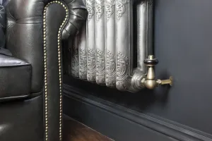Windsor Corner TRV in Natural Brass with a black head, fitted to an ornate silver cast iron radiator beside a leather Chesterfield sofa.