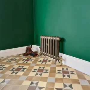 Low Emmeline I cast iron radiator in Beaten Brass on feet with brass valves, against a green wall above Victorian geometric floor tiles.