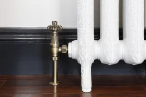 Chatsworth thermostatic valve in Antique Brass on an ornate white cast iron radiator on feet, against a dark skirting and wooden floor.