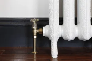 Chatsworth angled manual valve in Antique Brass fitted to an ornate white cast iron radiator on feet, dark skirting and wooden floor.