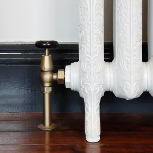 Windsor angled manual valve in Natural Brass with a black wheel head, fitted to an ornate white cast iron radiator foot on a dark timber floor.