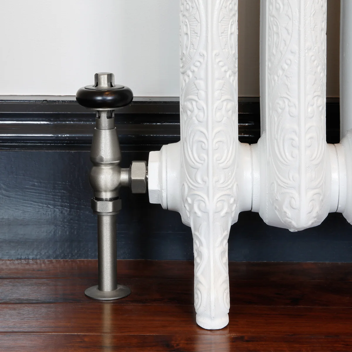 Windsor angled valve in Natural Pewter with a black wooden head, fitted to an ornate white cast iron radiator foot on a dark wood floor.
