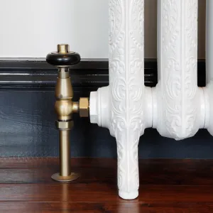 Windsor angled valve in Natural Brass with a black head, fitted to an ornate white cast iron radiator foot on a dark wooden floor.