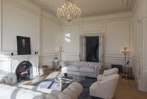 Low dark grey Grace 6 column cast iron radiator beside a doorway in a panelled drawing room with Chesterfield sofas and a chandelier.