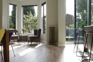 Tall Grace 4 Column cast iron radiator in Soft Pewter on a wall beside glazed doors, with a low radiator under a bay window and oak floor.