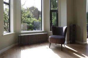 A Grace 6 Column 485mm cast iron radiator in Soft Pewter on feet under a bay window, with a tall radiator and grey armchair on an oak floor.