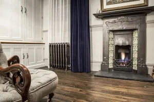 Ornate Rococo III cast iron radiator in Full Polish on feet by a window with navy velvet curtain, beside a tiled cast iron fireplace.