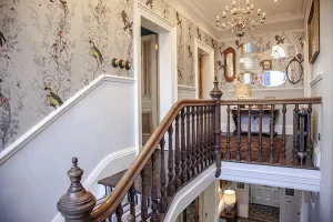 Ornate Rococo cast iron radiator in a dark finish, partly behind turned wooden banisters on a landing with bird wallpaper and a chandelier.